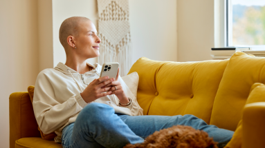 woman sitting up with legs across couch, phone in hand, and dog laying at bottom of her legs