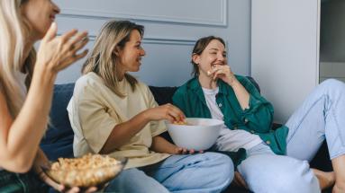 Group of 3 ladies sitting on couch eating popcorn and laughing 