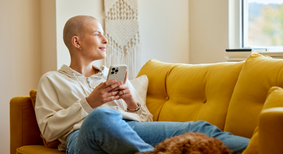 woman sitting up with legs across couch, phone in hand, and dog laying at bottom of her legs