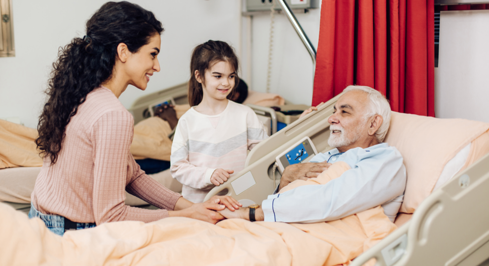 Man laying in hospital bed with a woman and female child by his bed side. 